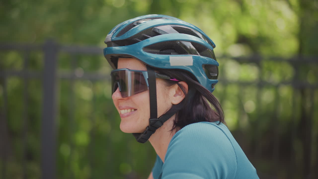 Mulher sorridente usando capacete de bicicleta e óculos de sol espelhados, rindo durante um passeio de lazer em um parque arborizado com uma camisa verde-azulado confortável, vegetação iluminada pelo sol, close-up espontâneo capturando um sorriso radiante, luz natural e aconchegante