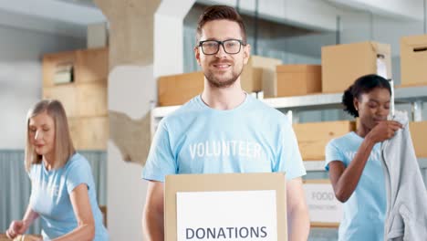 Caucasian-man-volunteer-holding-donation-boxes-and-looking-at-camera-in-charity-warehouse-while-his-coworkers-working-packing-boxes