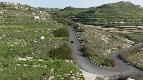 racing car driving fast on the road track up the lush hill on a bright sunny day - aerial shot