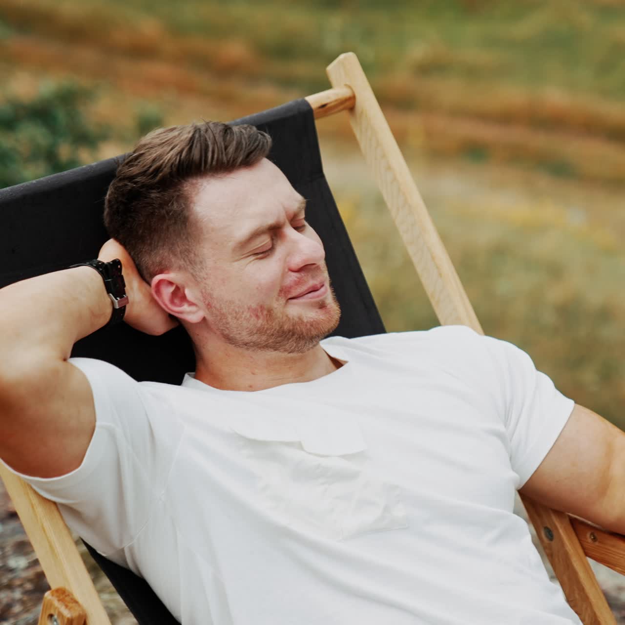 Relaxed man laid back in the folding chair outdoors. Man talk on the phone by video chat holding one hand behind his head. Blurred backdrop. Top view