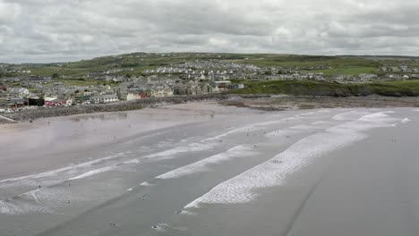 Aerial-retreats-from-wide-Lahinch-Beach,-surfers-on-small-shore-break