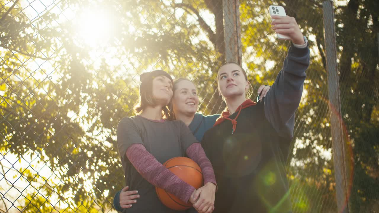 un trío de jugadoras de baloncesto de niñas felices en ropa deportiva toman una selfie juntos con una pelota de baloncestro en una cancha de balonceso al aire libre cerca de una rejilla en un día soleado