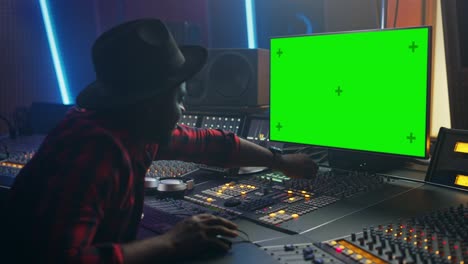 portrait of stylish audio producer working in music record studio, uses green screen computer, mixer board, control desk to create new song. creative black artist dances while sitting. over shoulder