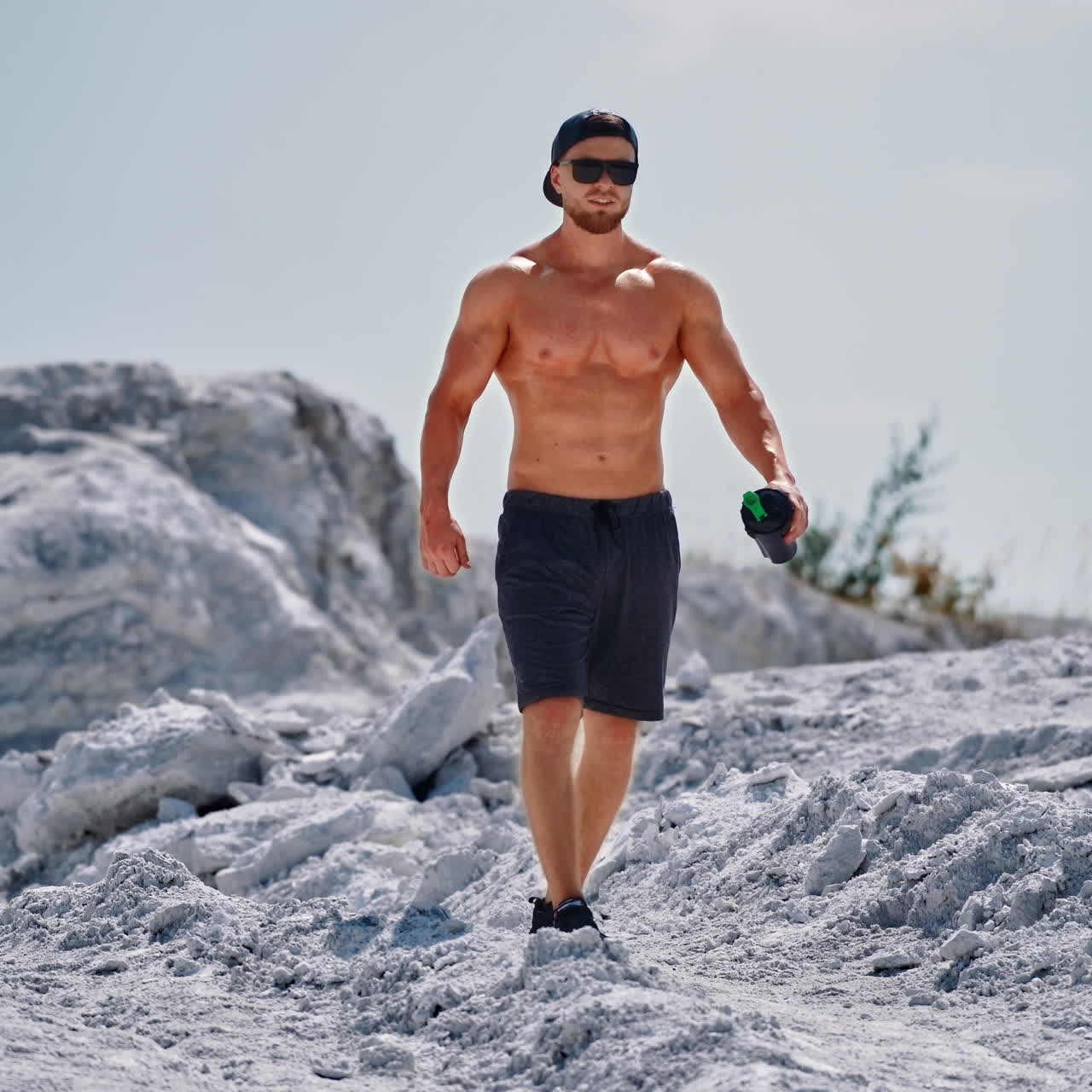 Handsome man with trained body holding flask in the mountains. Muscular athlete in shorts and sunglasses descends from the white hill in a sunny summer day.