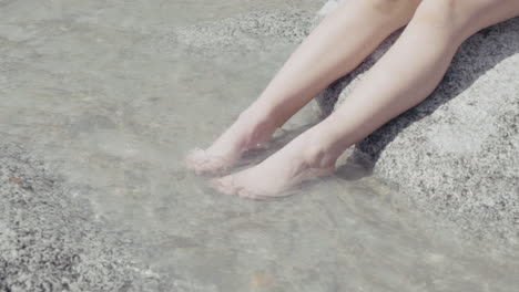 close-up of a little girl sitting on a stone and dipping her legs and feet in on stream on a sunny day in the italian alps