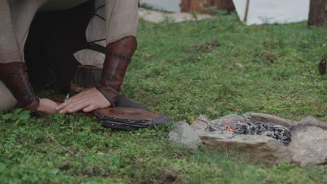 a person in historical reenactment clothing kneels on the grass, using a traditional hand-operated bellows to blow air onto the embers of a campfire, making them glow.