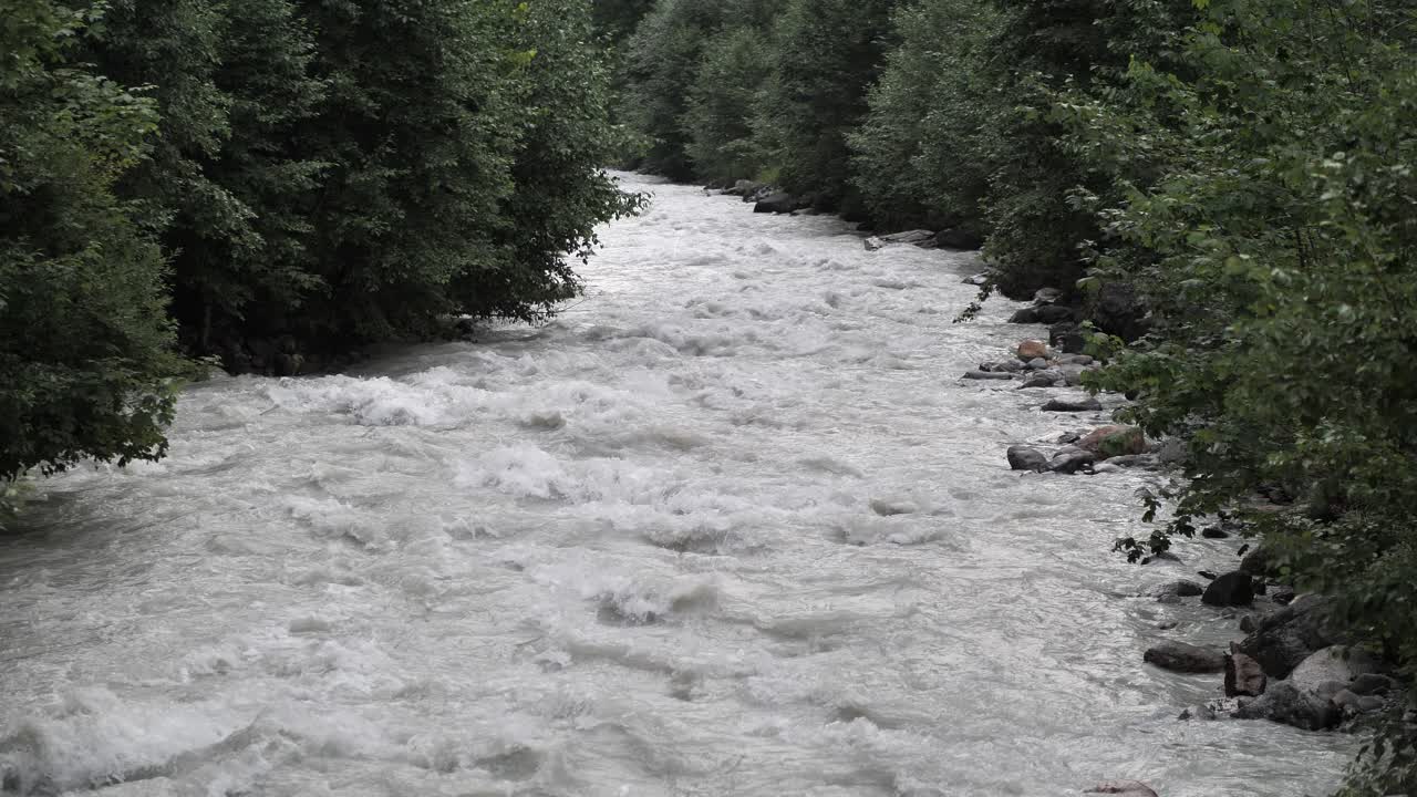 Lille flod af smeltet gråt vand, der strømmer og skvulper på klipperne, mens grønne træer vokser på siden. Vidvinkelbillede.