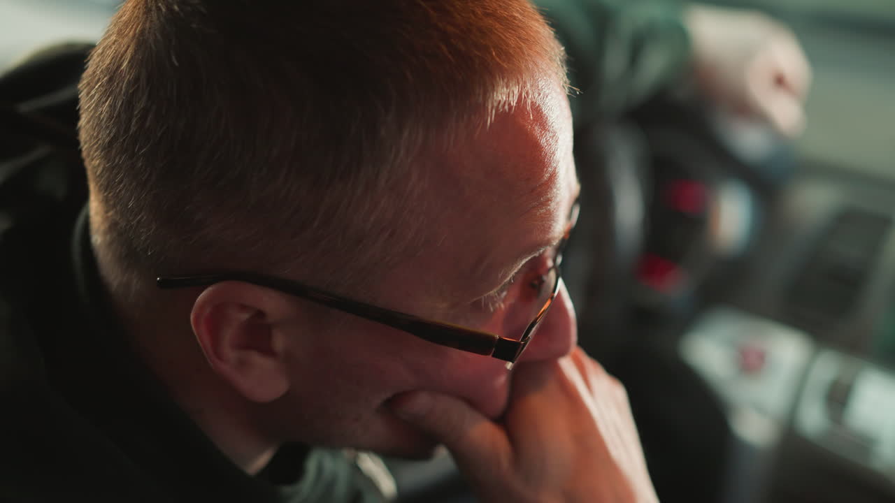A close-up side profile of a man deep in thought, sitting in a car, with his hand resting on his chin. The man is wearing glasses, and the interior of the car is visible