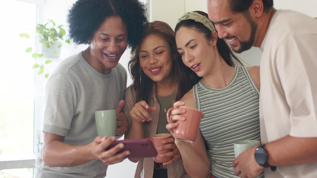 Amigos diversos reunidos en la cocina mientras un hombre muestra contenido en su teléfono, señalando y sosteniendo tazas.