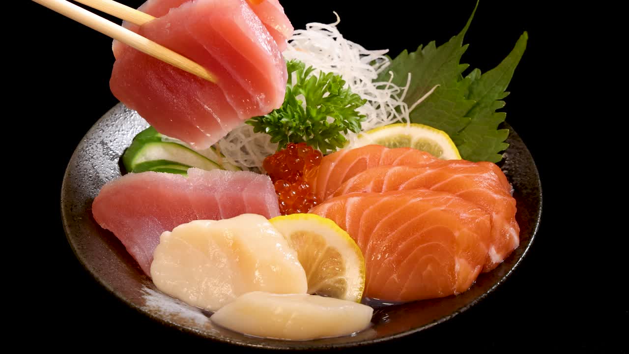 Chopsticks delicately lift a slice of tuna sashimi from a vibrant Japanese sushi platter, arranged on a black background with bright, even lighting