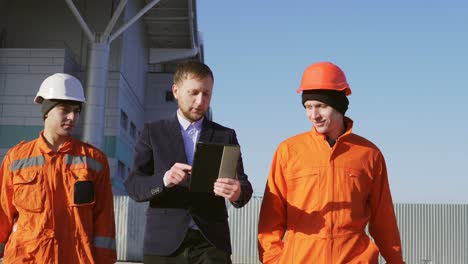 Un-Gerente-Con-Traje-Y-Dos-Trabajadores-Con-Uniforme-Naranja-Y-Cascos-Caminan-Por-Las-Instalaciones-Del-Edificio.-Filmado-En-4k