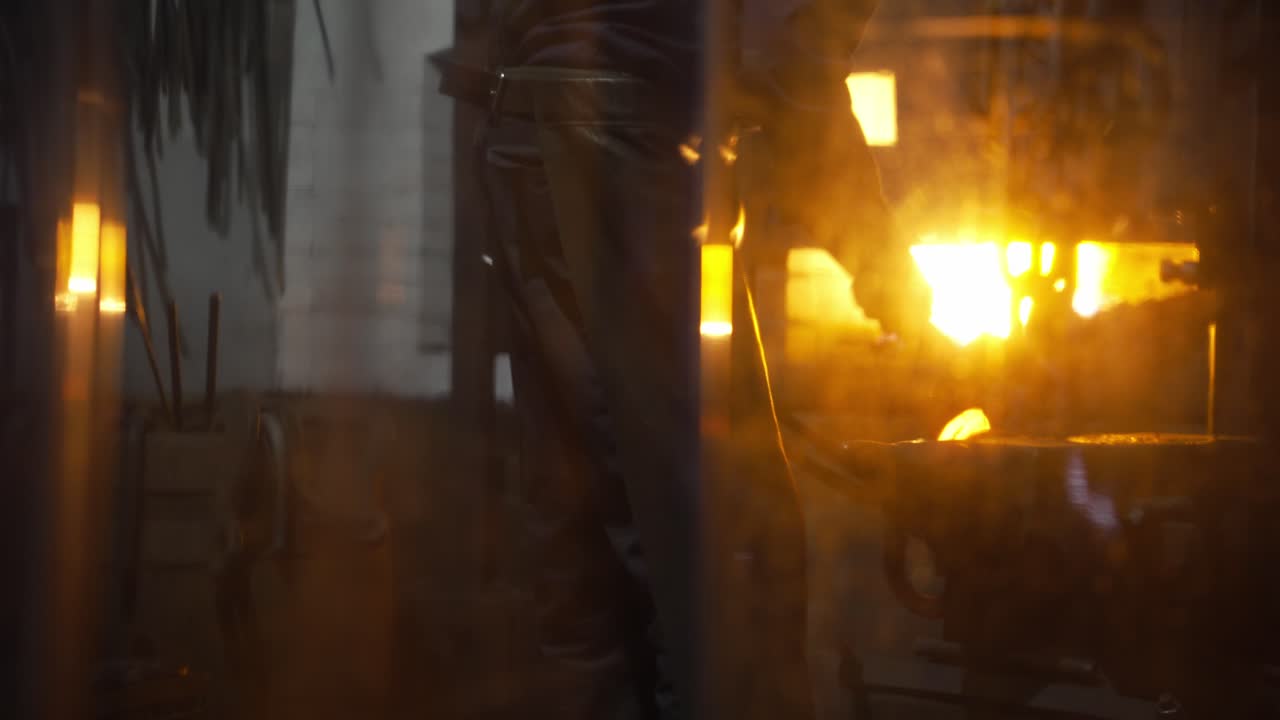 Blacksmith forging a heated chain link inside a forge workshop with strong warm backlight from the fire