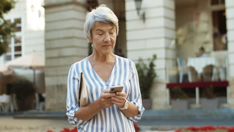 Hermosa-Anciana-De-Pie-En-El-Centro-De-La-Ciudad-Y-Tocando-El-Mensaje-En-El-Teléfono-Inteligente