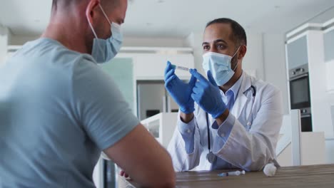 Mixed-race-male-doctor-wearing-face-mask-consulting-male-patient-at-home-before-vaccine
