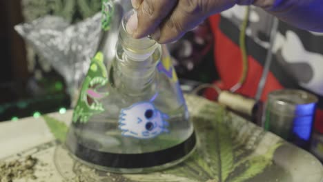una mano de un hombre negro recogiendo un brote de marihuana de una bandeja de rodadura de hojas de olla y empaquetándolo en el cuenco de un bong de agua de vidrio con un molinillo de hierba y una bolsa de brotes en el fondo