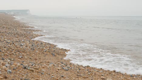 Slowmotion-shot-of-the-pebble-rocky-coast-of-Seaford-with-rocks-in-the-foreground-and-white-chalk-cliffs-in-the-background