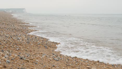 Toma-En-Cámara-Lenta-De-La-Costa-Rocosa-De-Guijarros-De-Seaford-Con-Rocas-En-Primer-Plano-Y-Acantilados-De-Tiza-Blanca-En-El-Fondo