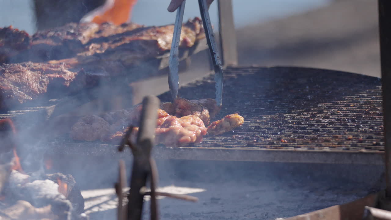 Man's hand with tongs arranging grilled meat asado, traditional Argentine food with burning and smoking firewood, close up