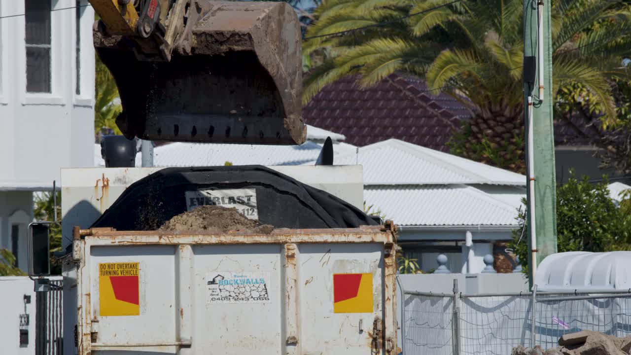 Excavator dumps soil into dump truck at urban construction site under bright daylight