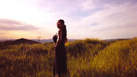 Mujer-Hispana-Vestida-De-Negro-Parada-En-Una-Pradera-Bajo-La-Espectacular-Luz-Del-Sol-De-La-Hora-Dorada-Y-Mirando-El-Horizonte