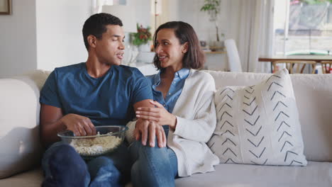 Una-Pareja-De-Adultos-Jóvenes-Afroamericanos-En-El-Sofá-De-Su-Sala-De-Estar-Hablando,-Riendo-Y-Comiendo-Palomitas-De-Maíz