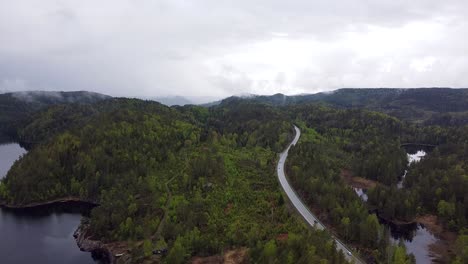 Aerial-view-of-a-winding-road-cutting-through-lush,-green-Norwegian-forest-with-misty-hills-in-the-background