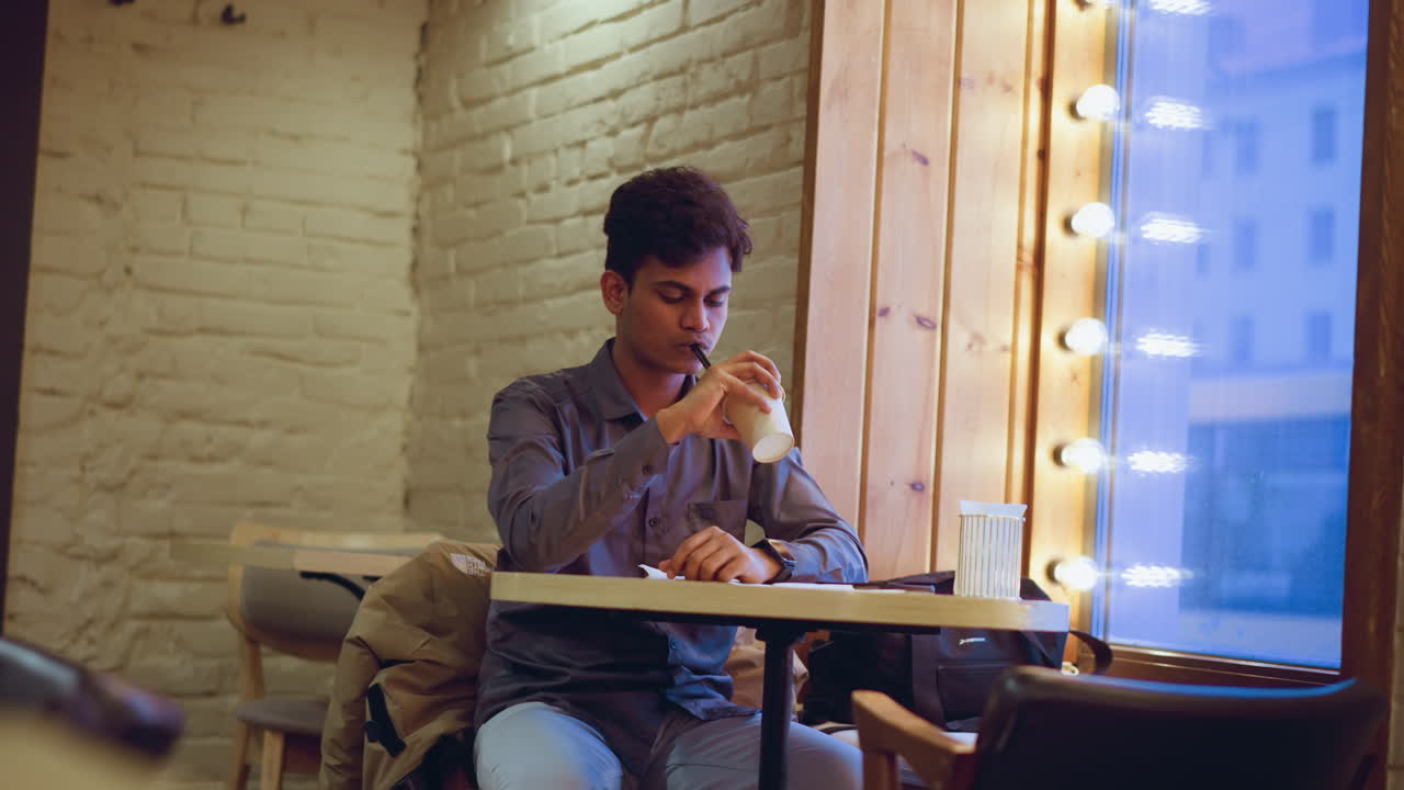 Jovem de camisa cinza sentado sozinho perto de uma grande janela com luzes de corda em um café moderno e aconchegante, segurando uma xícara de café e um canudo, parecendo pensativo e relaxado em uma atmosfera interna calma e suavemente iluminada.