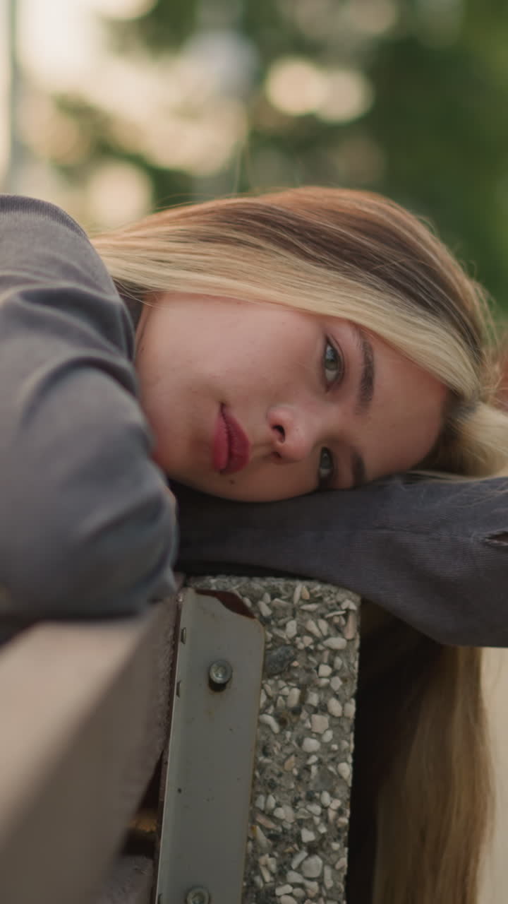Close-up of woman in grey clothing resting head on arm outdoors, lost in thought with a calm expression, background is blurred featuring greenery
