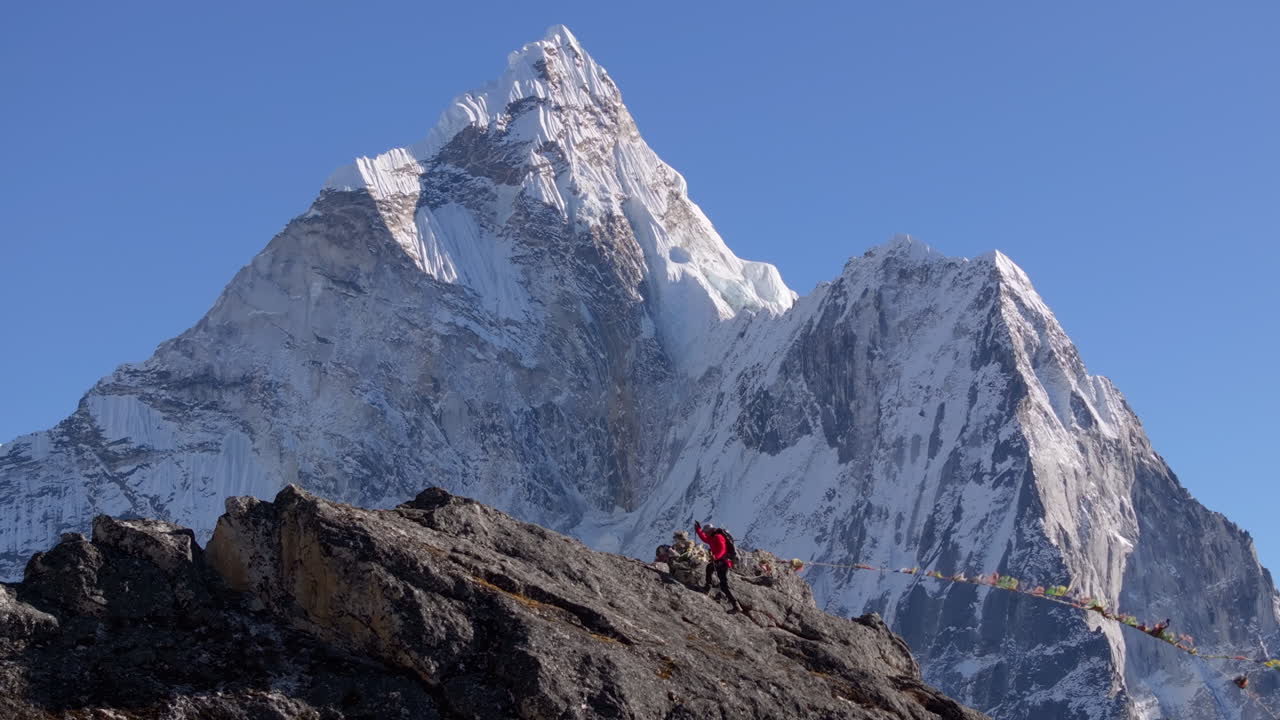 Ripresa con teleobiettivo da drone di un escursionista in ascesa verso la vetta Nangkartshang sopra Dingboche lungo il percorso del Campo Base dell'Everest in Nepal. L'Ama Dablam si erge sullo sfondo mentre le bandiere di preghiera sventolano al vento.