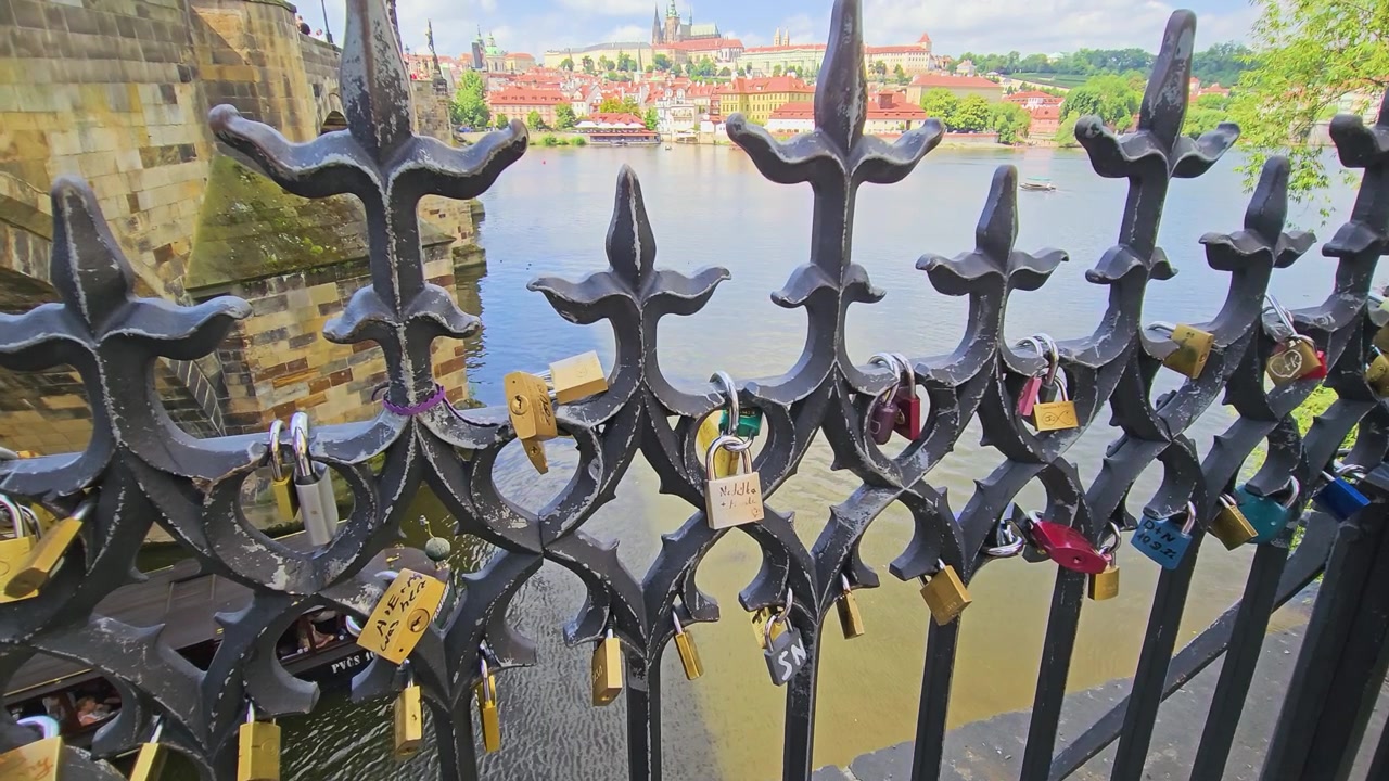 Primo piano di lucchetti d'amore appesi a una recinzione in ferro ornata vicino al Ponte Carlo a Praga, con il fiume Moldava, il vecchio ponte in pietra, i tetti rossi e il Castello di Praga sfocati sullo sfondo