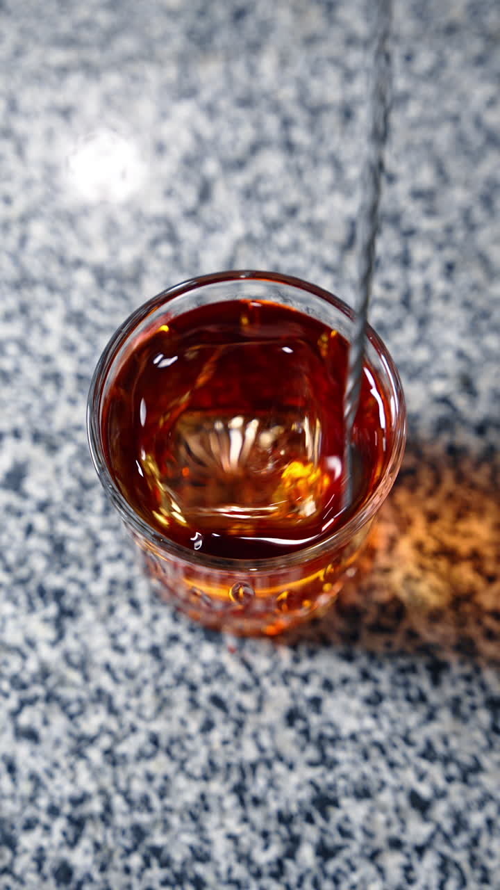 Brown drink with a big ice cube mixed with a long spoon. Preparation of a beverage in the bar. Top view. Vertical video.