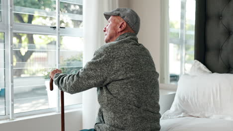 Elderly man sitting on bed looking out window with cane