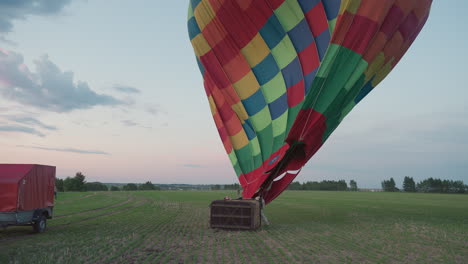 person guiding deflating colorful hot air balloon canopy across expansive green farmland while pulling tether rope toward waiting red equipment trailer under pastel dusk sky