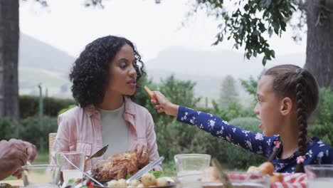 happy biracial mother and daughter eating meal at dinner table in garden, slow motion