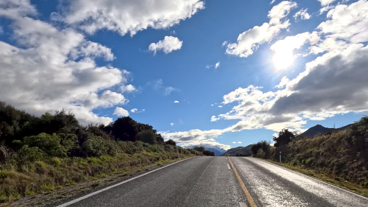 Carro percorre uma estrada sinuosa através de uma paisagem exuberante, montanhas e nuvens dramáticas sob a luz forte do sol
