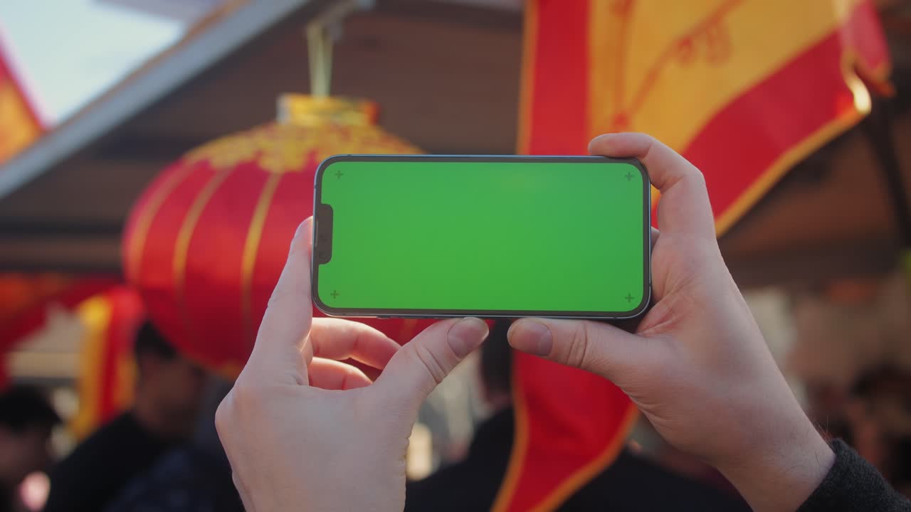 Young man holding phone chroma key screen on the street in front of chinese lignt close-up. Male with green screen on smart phone swiping .