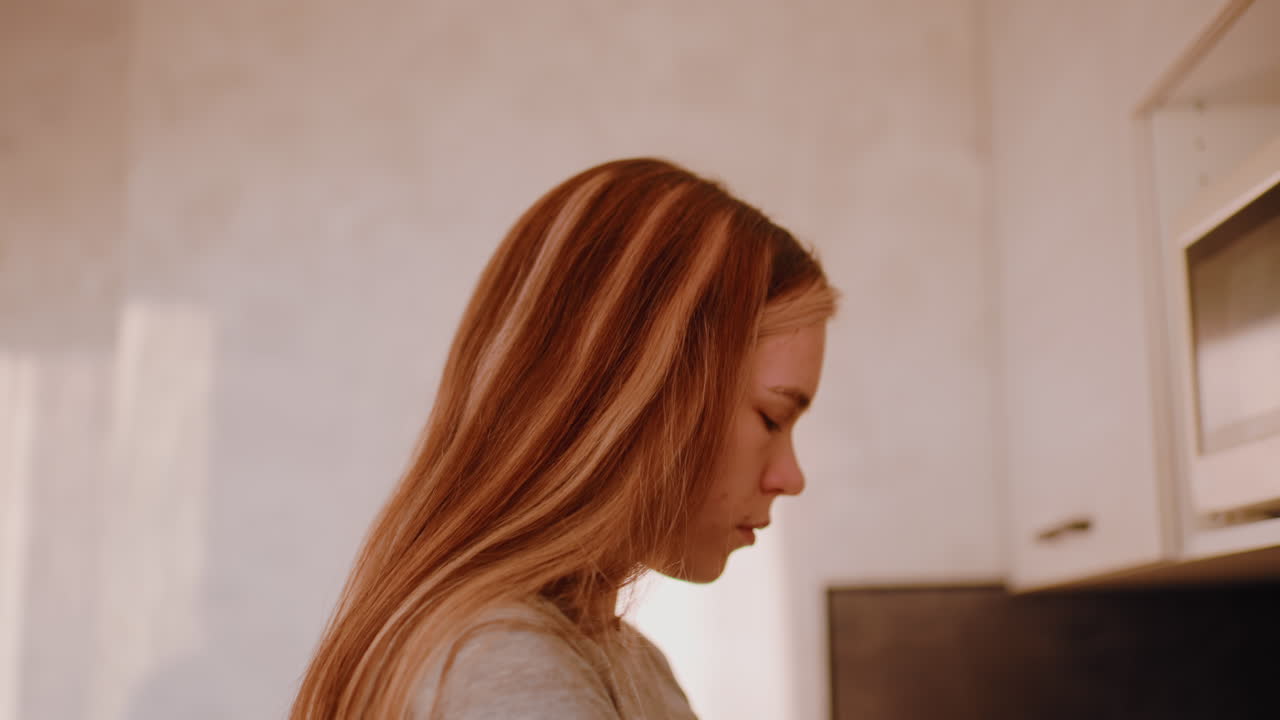 Close up of young woman in casual grey shirt retrieving item from kitchen cabinet with natural daylight softly illuminating her face as she prepares to start morning routine