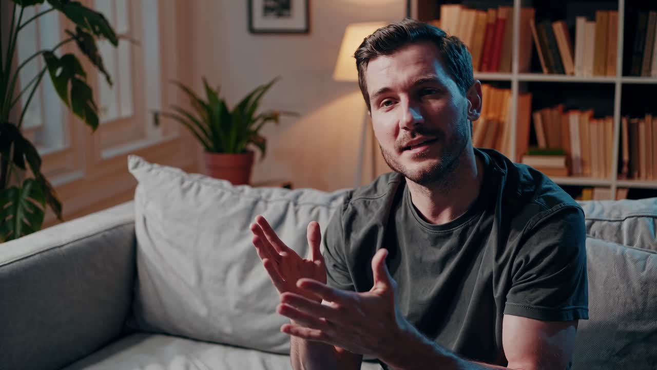 En video i halvfigur av en man i en soffa som gestikulerar medan han pratar. Varm belysning och en mysig miljö.