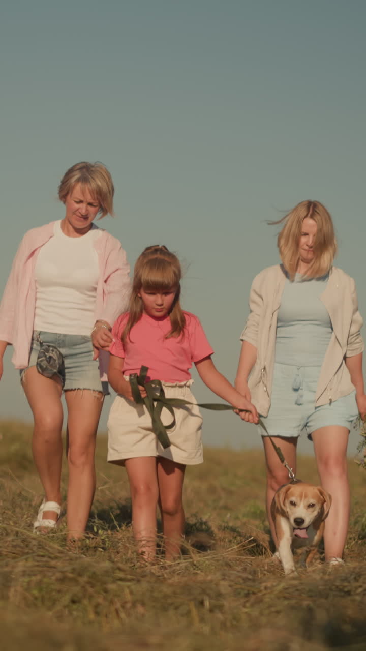 две женщины идут рядом с маленьким ребенком, держащим собачью повязку, в то время как собака останавливается, чтобы посмотреть на женщину в розовом, женщина в синем держит букет цветов, сцена передает семейную связь