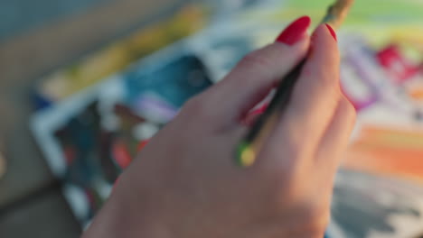 manicured woman hands applying vibrant brushstrokes on blurred paper full of mixed colors on wooden table outdoors at sunset captured, focus highlighting texture motion and creative process
