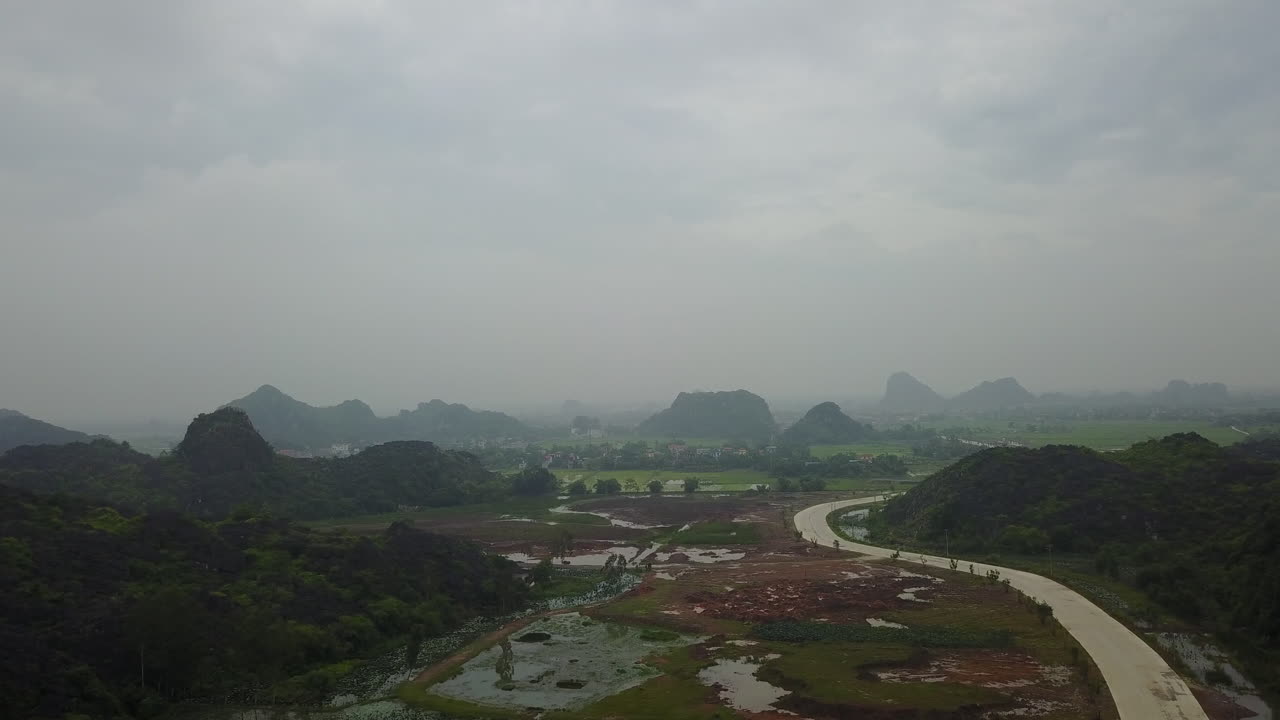A scenic view of a road winding through the karst rock formations in Ninh Binh, Vietnam, on a cloudy day. The dramatic limestone peaks and lush greenery create a breathtaking landscape.