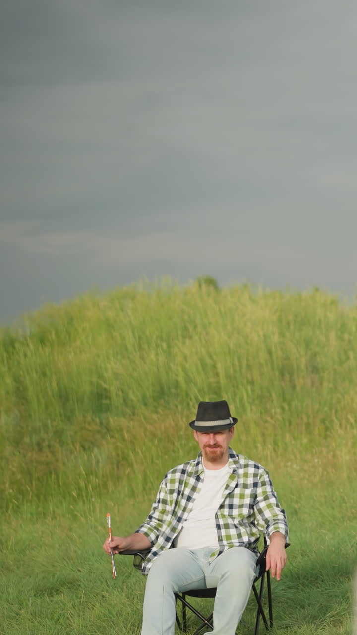 un hombre con un sombrero negro, camisa a cuadros y vaqueros se sienta cómodamente en una silla en un campo cubierto de hierba iluminado por el sol. el ambiente sereno y la vestimenta casual sugieren un momento de paz en la naturaleza