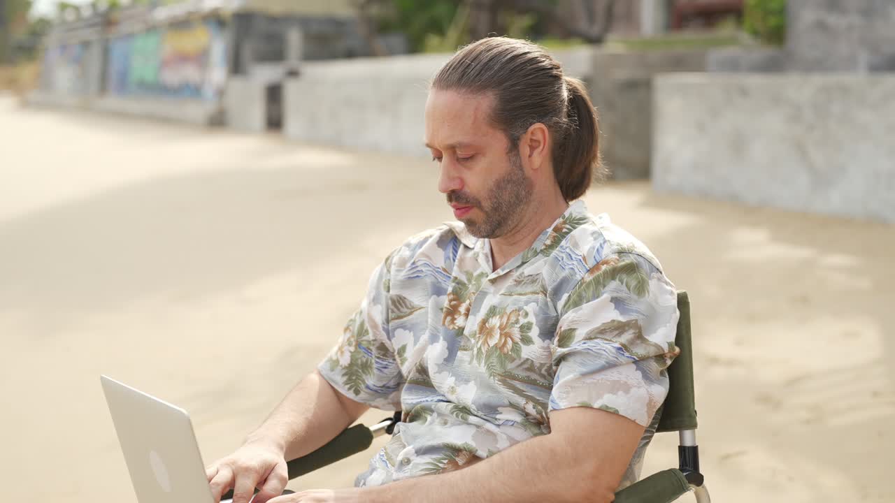 empresario caucásico de 4k sentado en una silla al aire libre y trabajando en una computadora portátil en la playa.