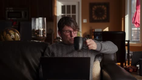 older man drinking coffee while sitting on a couch and working at home