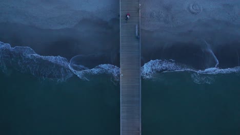 top down drone footage of a pier with a fisherman walking and waves crashing