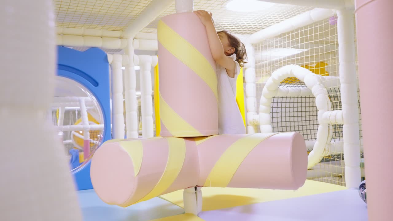 Happy little girl laughs with joy while riding a soft marshmallow-themed spinning obstacle at a modern indoor playground and entertainment center for kids