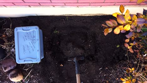 Digging-a-hole-and-dispersing-dirt-from-the-ground-of-a-flower-garden-while-the-plastic-case-with-a-dead-family-pet-lays-waiting-to-be-buried-in-slow-motion