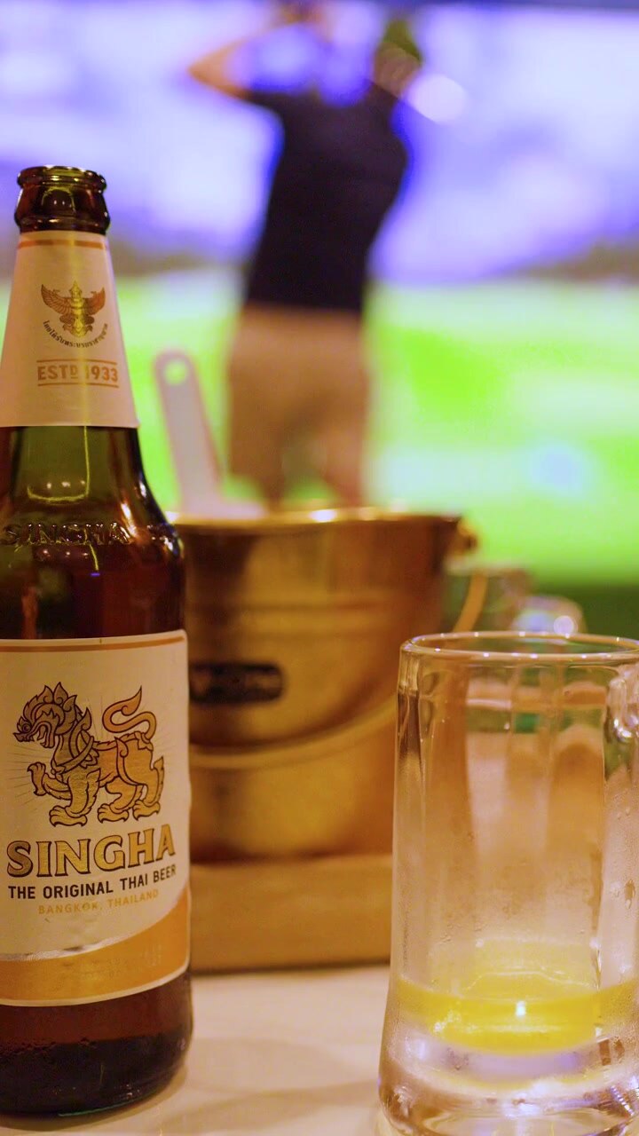 Una bottiglia di birra e un bicchiere freddi sono in primo piano mentre un uomo pratica golf indoor.