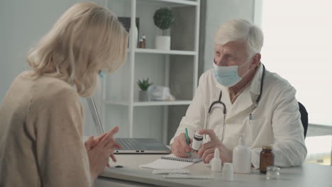 a grey haired, middle aged doctor in a facemask prescribes medicines to a female patient 1