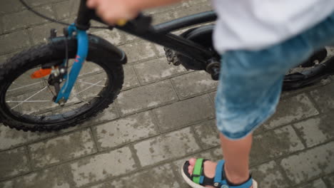 a lower view of a child standing beside a bicycle, carefully rides the bicycle, the child wears denim shorts and colorful sandals, and the bicycle features a blue front wheel